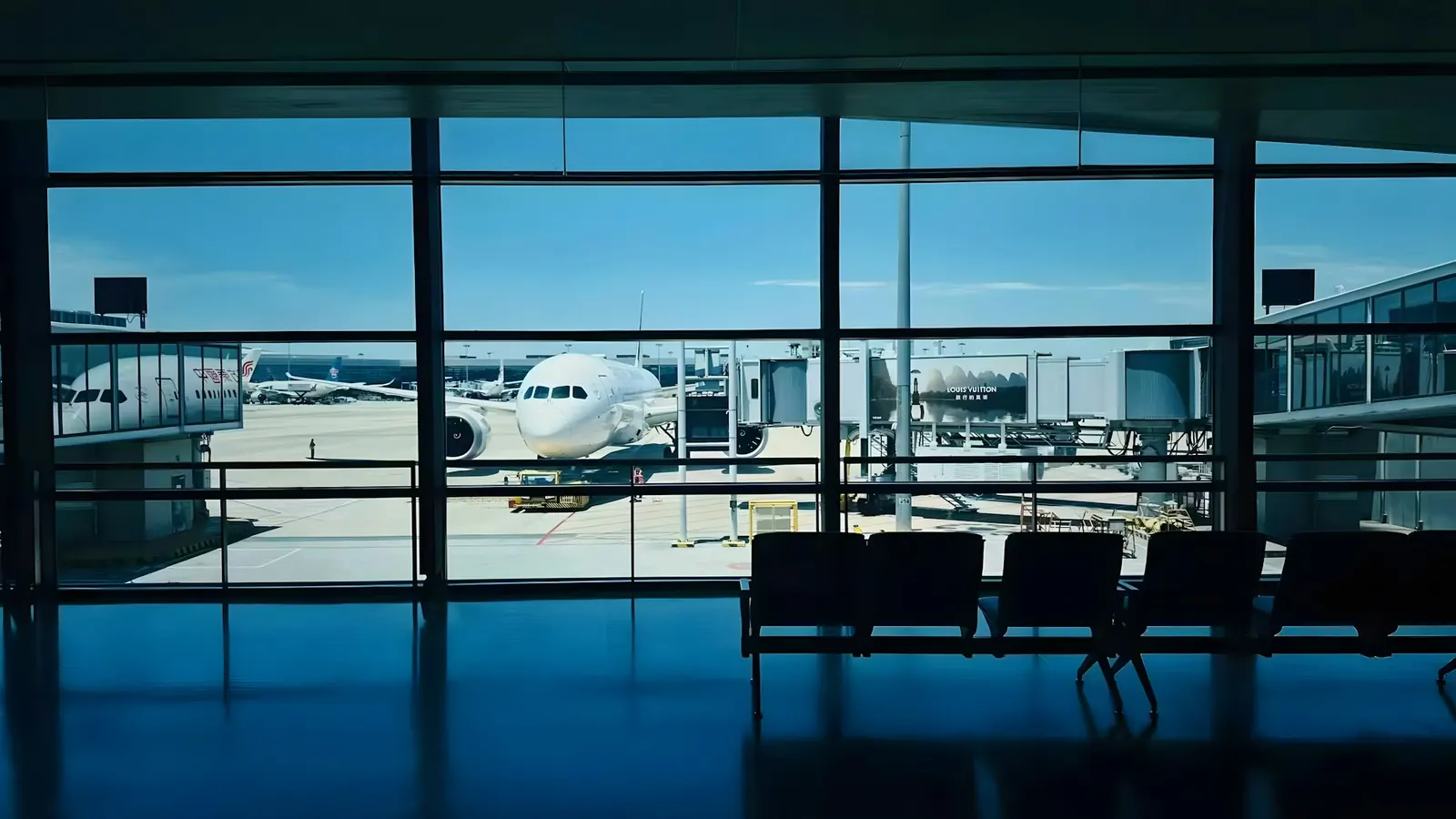 Blick aus einem Flughafenterminal durch große Glasfenster auf ein Flugzeug am Gate, davor leere Sitzbänke